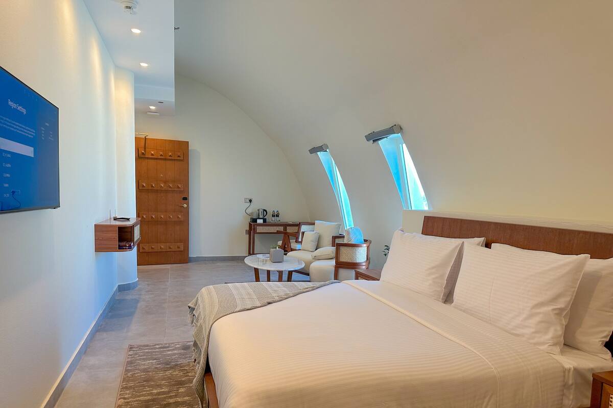 A modern bedroom with a large bed, a small seating area, and a wooden wardrobe.