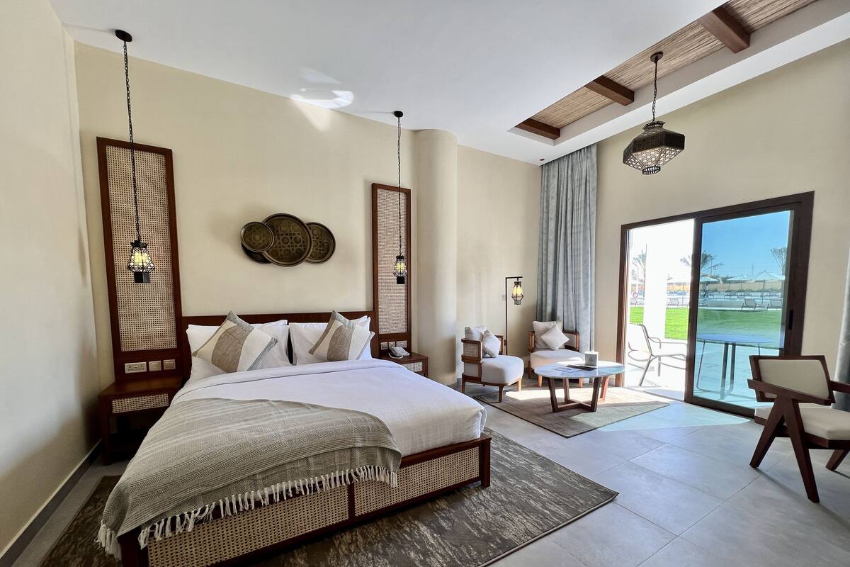 A spacious bedroom with a large bed, two armchairs, a small table, and a balcony with a view.