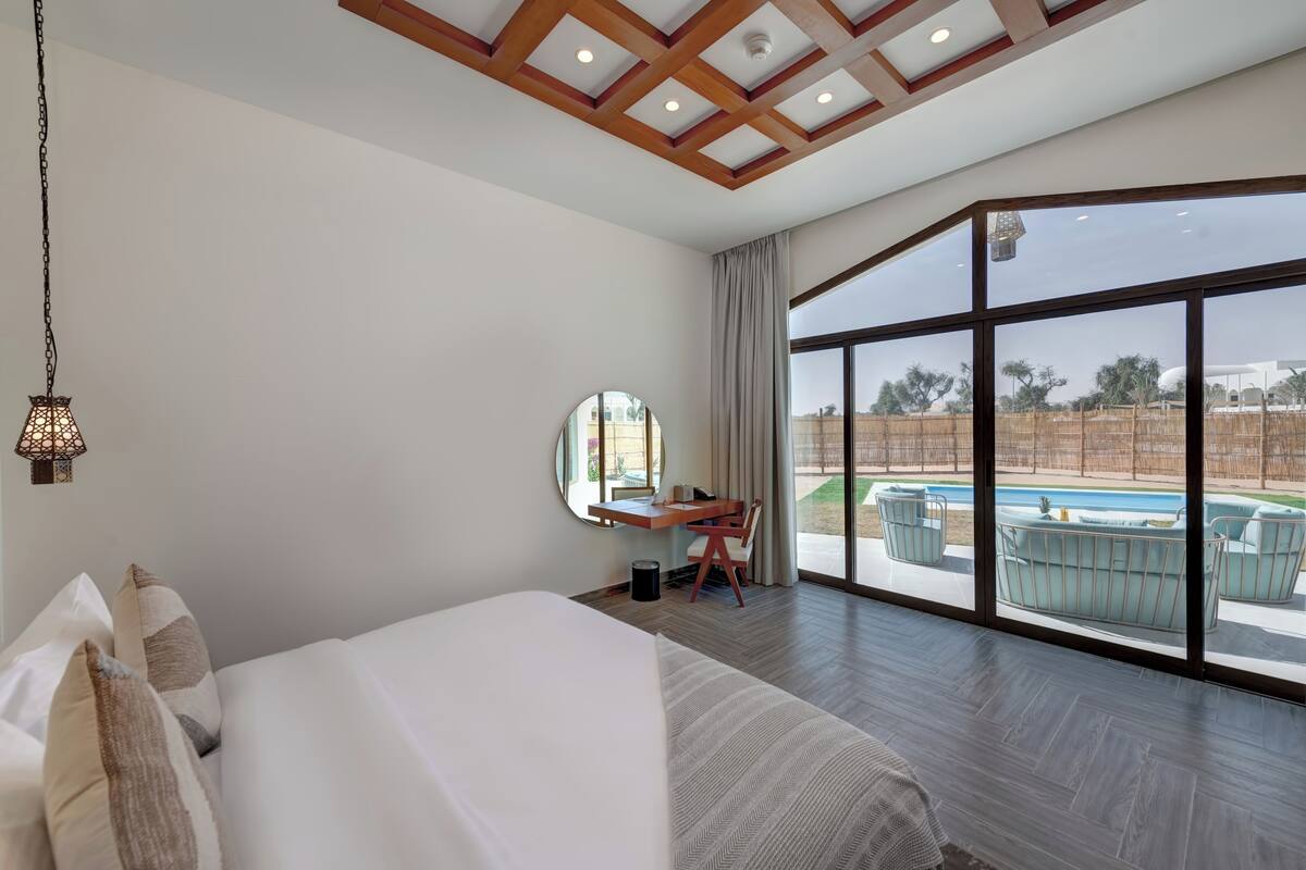 A hotel room with a large bed, a desk with a chair, a round mirror, and a view of an outdoor swimming pool.
