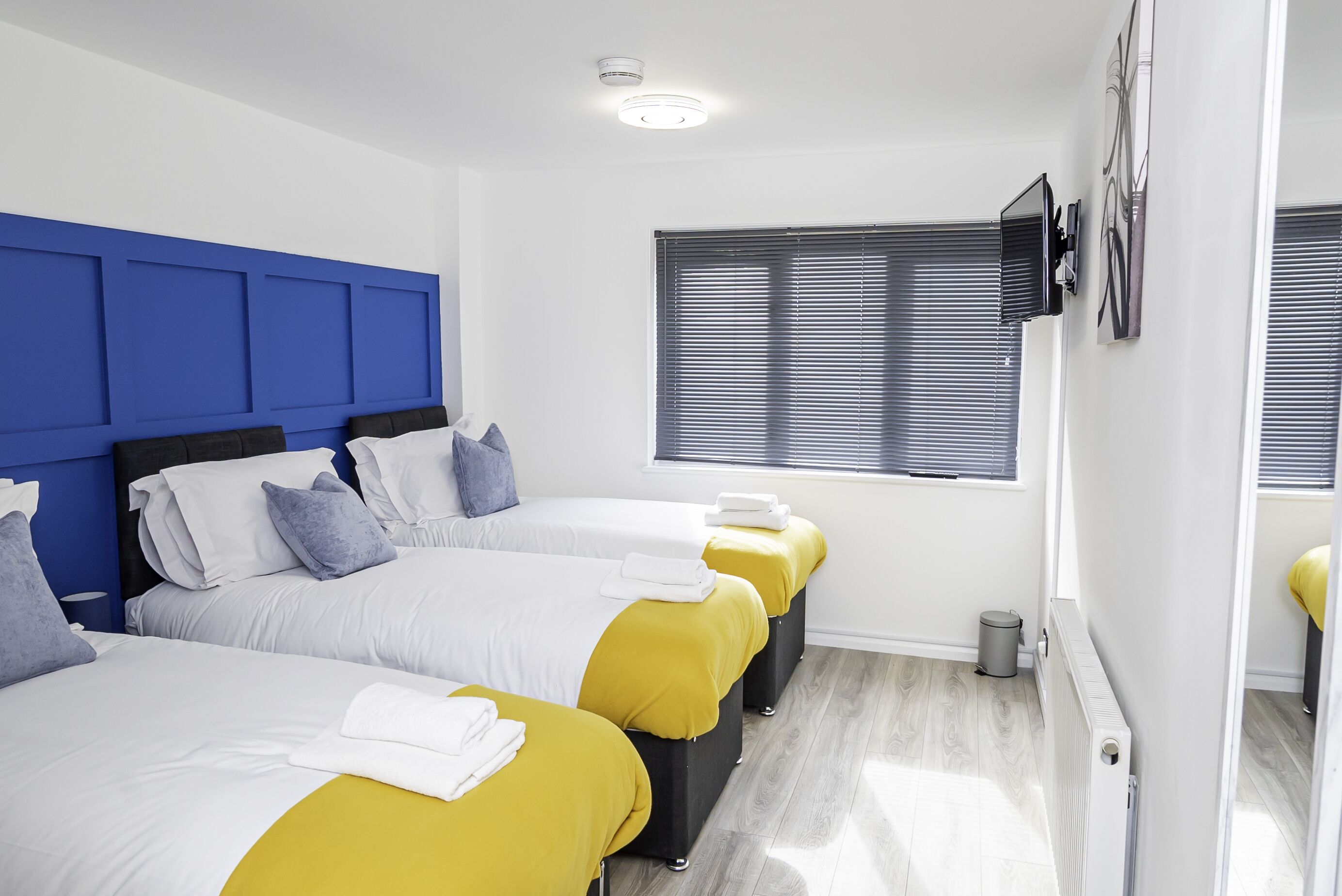 A hotel room with three beds, each with a different colored bedspread (white, yellow, and blue), and a large window with blinds.