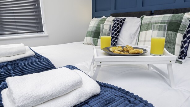 Un lit bien fait, orné d’un oreiller à carreaux bleus et blancs, une table basse avec une assiette de nourriture et deux verres de jus d’orange, et une fenêtre dont les stores sont fermés.