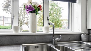 Une cuisine dotée d’un évier en acier inoxydable, d’une fenêtre donnant sur des arbres et des maisons, et d’un vase rempli de fleurs posé sur le rebord de la fenêtre.