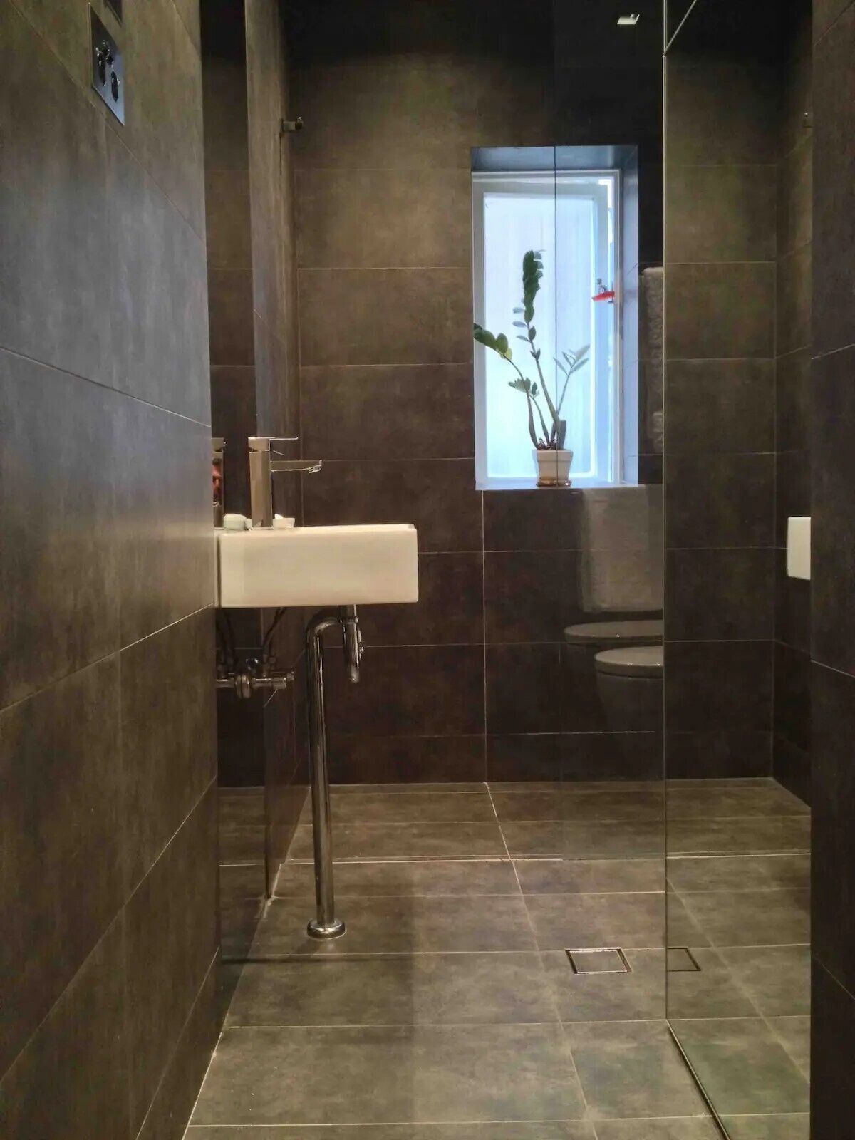 Un baño moderno con azulejos oscuros, un lavamanos blanco y una ventana con una planta.