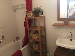 Ett badrum med en hylla i trä där korgar och en krukväxt står, en röd duschdraperi, ett vitt handfat och en spegel ovanför handfatet.