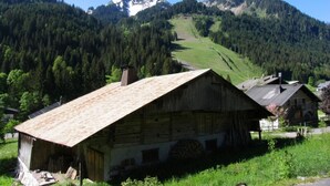 Una cabaña rústica de madera con techo inclinado, rodeada de exuberante vegetación y montañas al fondo.