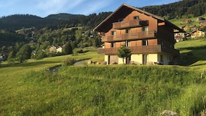 En storslagen chalet i trä med flera våningar och balkonger, belägen i en frodig, grön dal med ett berg i bakgrunden.