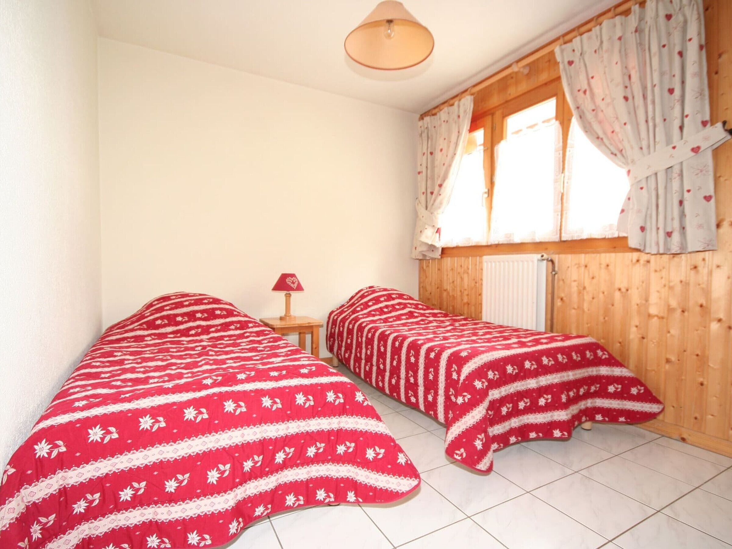 A small room with two beds, a window with curtains, a lamp, and a radiator.