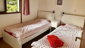 Un petit logement composé d’une seule pièce, avec deux lits, une fenêtre donnant sur un espace verdoyant, une table de chevet et un tableau accroché au mur.