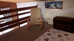 A room with a wooden ceiling, a chair with a heart-patterned cushion, a bed with a patterned blanket, a wooden dresser, and a mountain landscape painting on the wall.