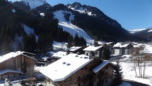 A snow-covered mountain village with chalet-style buildings, a ski slope in the background, and parked cars.