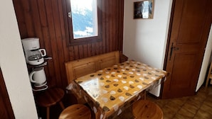 Ett mysigt rum med ett träbord, två pallar, en kaffemaskin, ett fönster med utsikt över snön och en dörr.