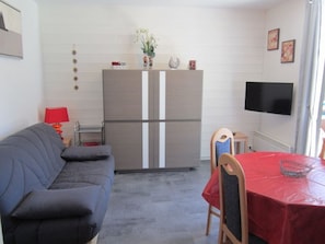 Un espacio habitable compacto con un sofá gris, una pequeña mesa de comedor con mantel rojo, un televisor montado en la pared y un refrigerador.