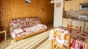 Een gezellige kamer met een houten vloer, een bank met een gestreept hoesje, een eettafel met een tafelkleed, houten kasten en een magnetron.