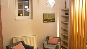 Une pièce comprenant deux fauteuils, une petite table, un radiateur, une fenêtre avec des stores, un luminaire suspendu et une cloison en bois dotée d’étagères.