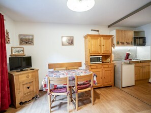 A compact room with a bed, a small table with a quilt, a TV on a wooden cabinet, a microwave, a sink, and wooden cabinets.