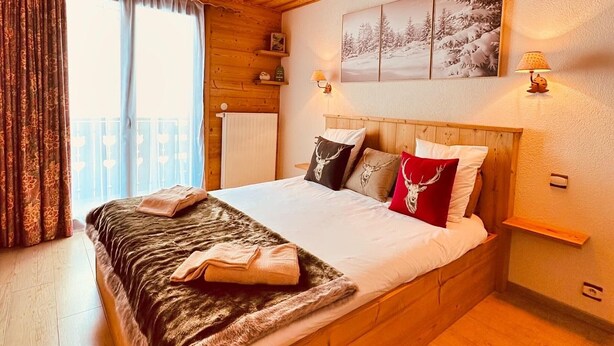 A wooden-framed bed with a fur throw, a red and white deer head pillow, and a black and white deer head pillow. There is a wooden shelf on the wall, a window with curtains, and a wall-mounted lamp. - Chalet in Châtel 6 bedrooms, 12 people (Châtel)