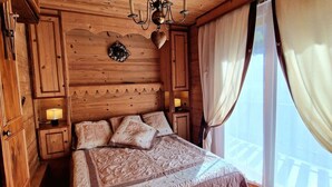 A wooden cabin-style room with a bed, built-in cabinets, a chandelier, and a large window with curtains. - Charming 2-BR Apt for 6 with Balcony, Near Super-Châtel Lift (Châtel)