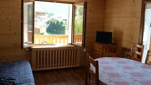 Une pièce de cabane en bois comprenant un lit, une table, un téléviseur et une fenêtre donnant sur des maisons et des arbres.