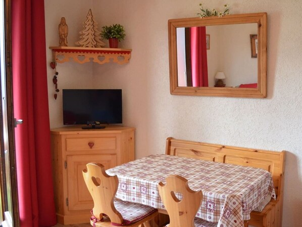 Une pièce avec un lit en bois, une table recouverte d’une nappe à carreaux, un téléviseur posé sur un meuble en bois, un miroir au mur et un rideau rouge.