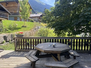 A wooden deck with a round table and benches, overlooking a mountainous landscape with houses and greenery.