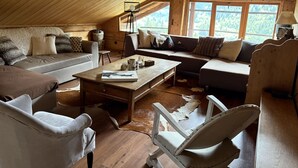 A cozy loft-style living room with a high ceiling, wooden beams, and large windows offering a view of a forested area. - 4-Room Chalet, 8 People, Wifi, Parking, Châtel (Châtel)