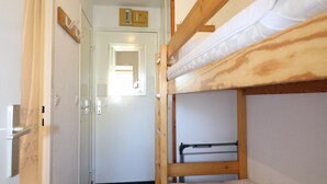 A narrow corridor leading to a door with a small window, bunk beds with wooden frames, and a wall-mounted control panel.