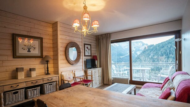 A cozy room with a wooden dining table, chairs, a sofa, a TV, and a large window offering a mountain view. - 3-room apartment for 6 people with balcony in Châtel (Châtel)