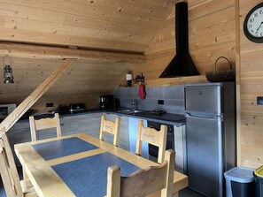 A wooden interior with a kitchen area, including a dining table, chairs, a refrigerator, and a clock on the wall.
