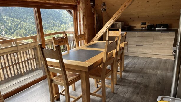 Puinen ruokailutila, jossa on pöytä ja tuolit, ja suurista ikkunoista avautuu näkymä metsään. Lisäksi tilassa on keittiö, jossa on kodinkoneet.