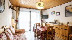 A cozy room with a wooden ceiling, a dining table with chairs, a sofa, a cabinet with a television, and a large window with curtains. - Cozy 2-Room Apartment for 5 Close to Village & Lifts, Pet-Friendly (Châtel)