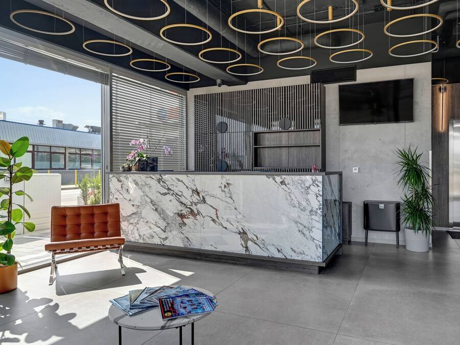 A modern lobby with a marble reception desk, a seating area with a brown chair, and a large TV screen.