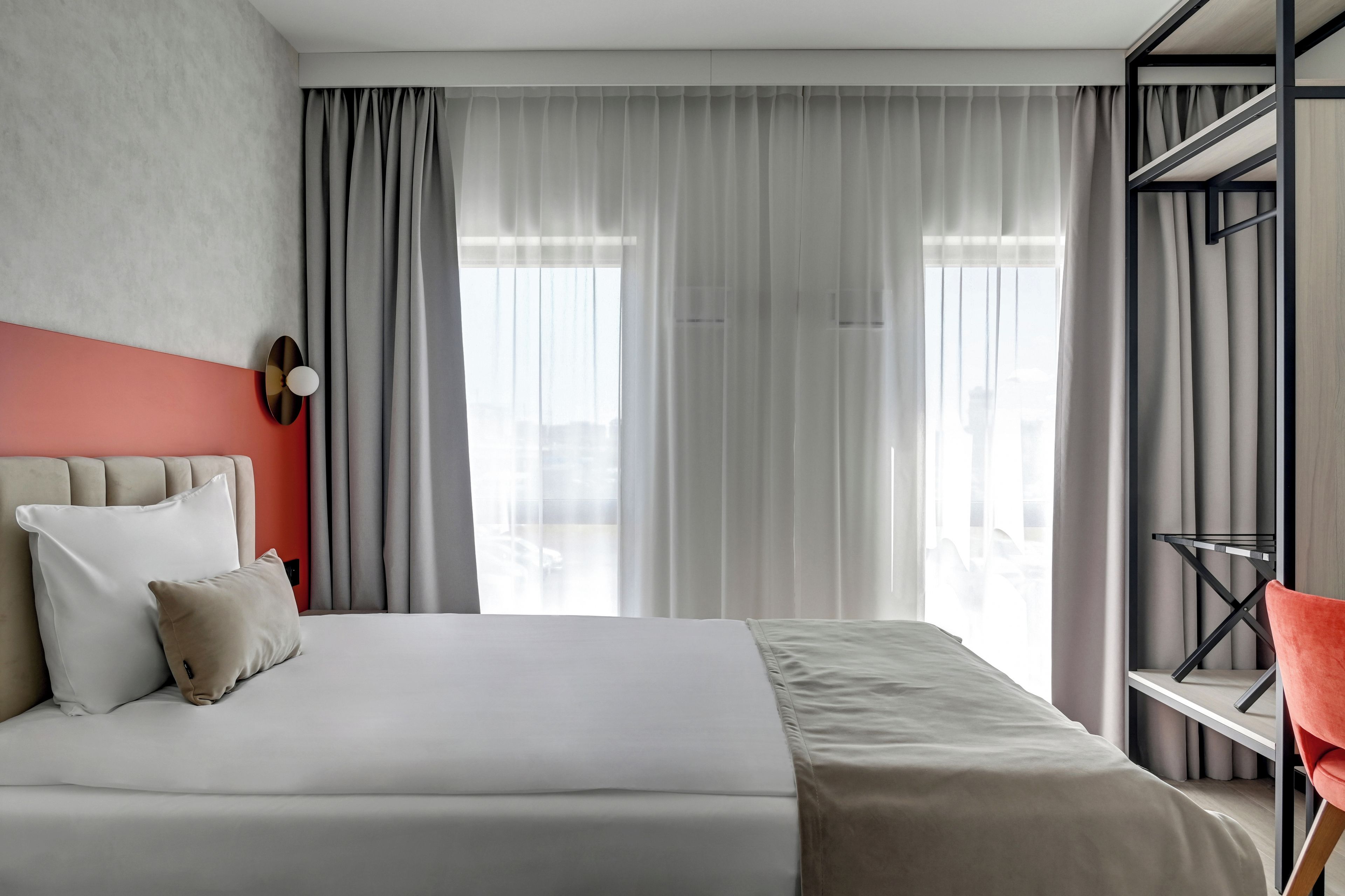 Une chambre d’hôtel moderne, dotée d’un lit, d’un mur d’accent rouge et offrant une vue sur la ville grâce à de grandes fenêtres.