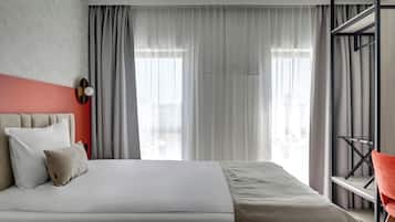 Une chambre d’hôtel moderne, dotée d’un lit, d’un mur d’accent rouge et offrant une vue sur la ville grâce à de grandes fenêtres.