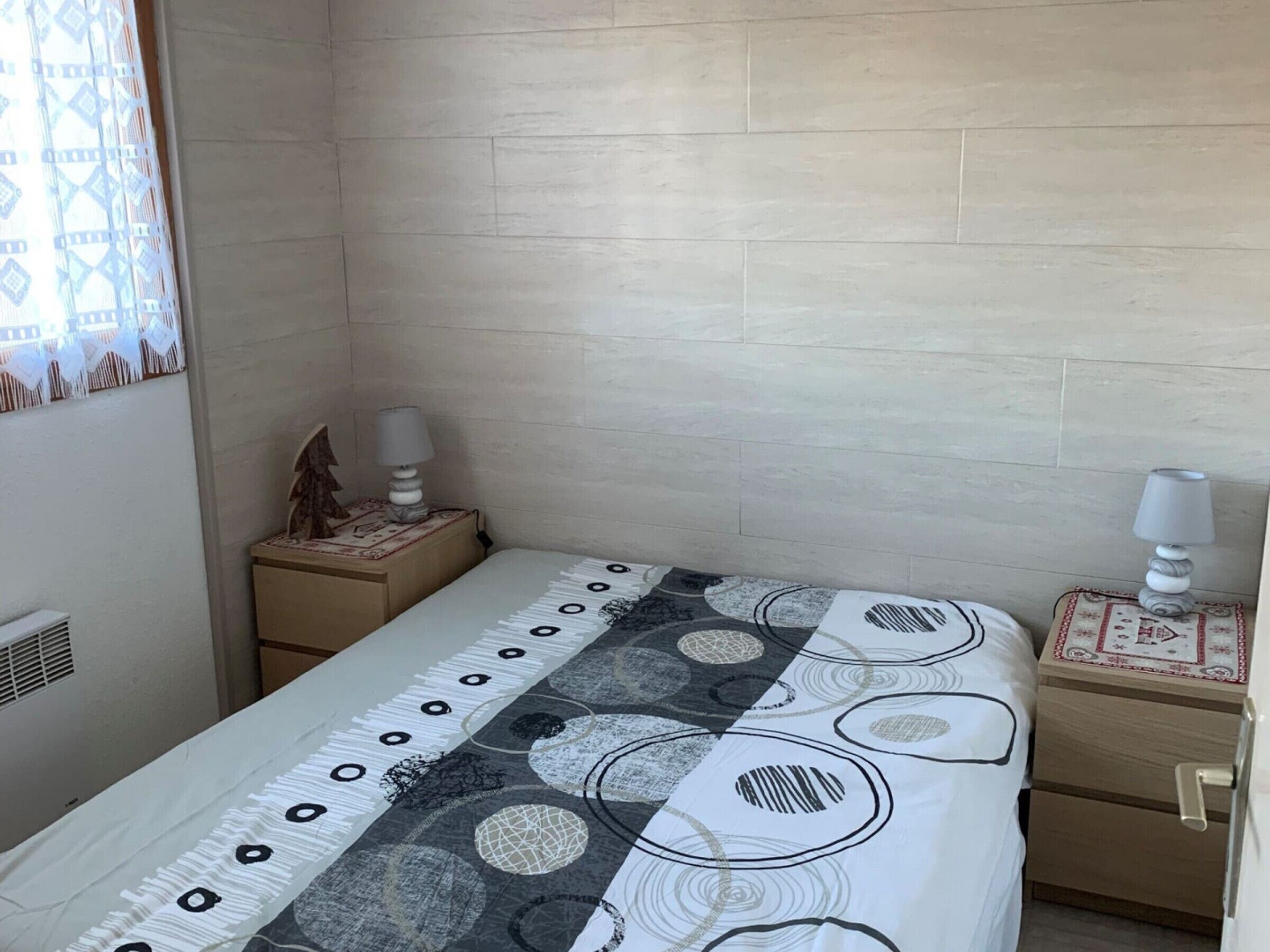 Une chambre à coucher comprenant un lit, deux tables de chevet, une commode, un radiateur fixé au mur et une fenêtre agrémentée de rideaux à motifs.