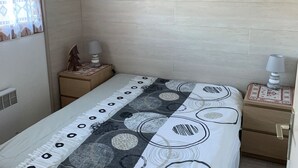 A bedroom with a bed, two bedside tables, a nightstand, a wall-mounted heater, and a window with patterned curtains. - Charming 2-Room Apartment, Sleeps 5, Terrace, Parking, Pet-Friendly (Châtel)