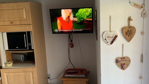 A television mounted on a wall, a wooden cabinet with a microwave, a small wooden table with a red object, and heart-shaped decorations on the wall. - Charming 2-Room Apartment, Sleeps 5, Terrace, Parking, Pet-Friendly (Châtel)