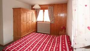 Ein Schlafzimmer mit einem Holzschrank, einem Heizkörper, einem Bett mit rot-weißer Bettdecke und einem Fenster mit Vorhängen.