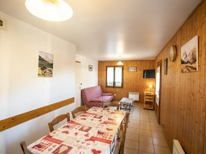 A cozy dining area with a long table, chairs, a patterned tablecloth, and wooden walls.