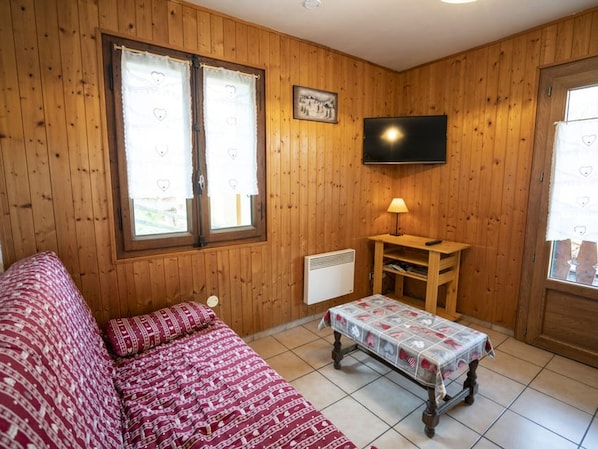 A cozy room with wooden walls, a bed with patterned bedding, a small table with a tablecloth, a heater, and a TV on the wall.