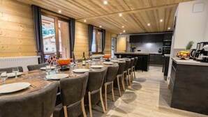 A modern dining area with a long wooden table, grey upholstered chairs, and a kitchen area with a fruit bowl and appliances. - Large 7-room chalet, 15 people, sauna, jacuzzi in Châtel (Châtel)