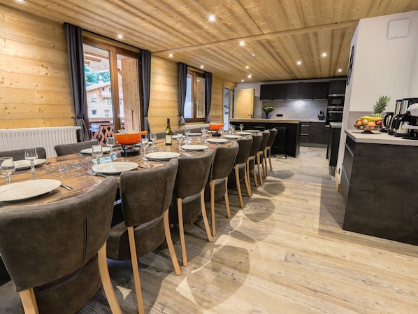 A modern dining area with a long wooden table, grey upholstered chairs, and a kitchen area with a fruit bowl and appliances.