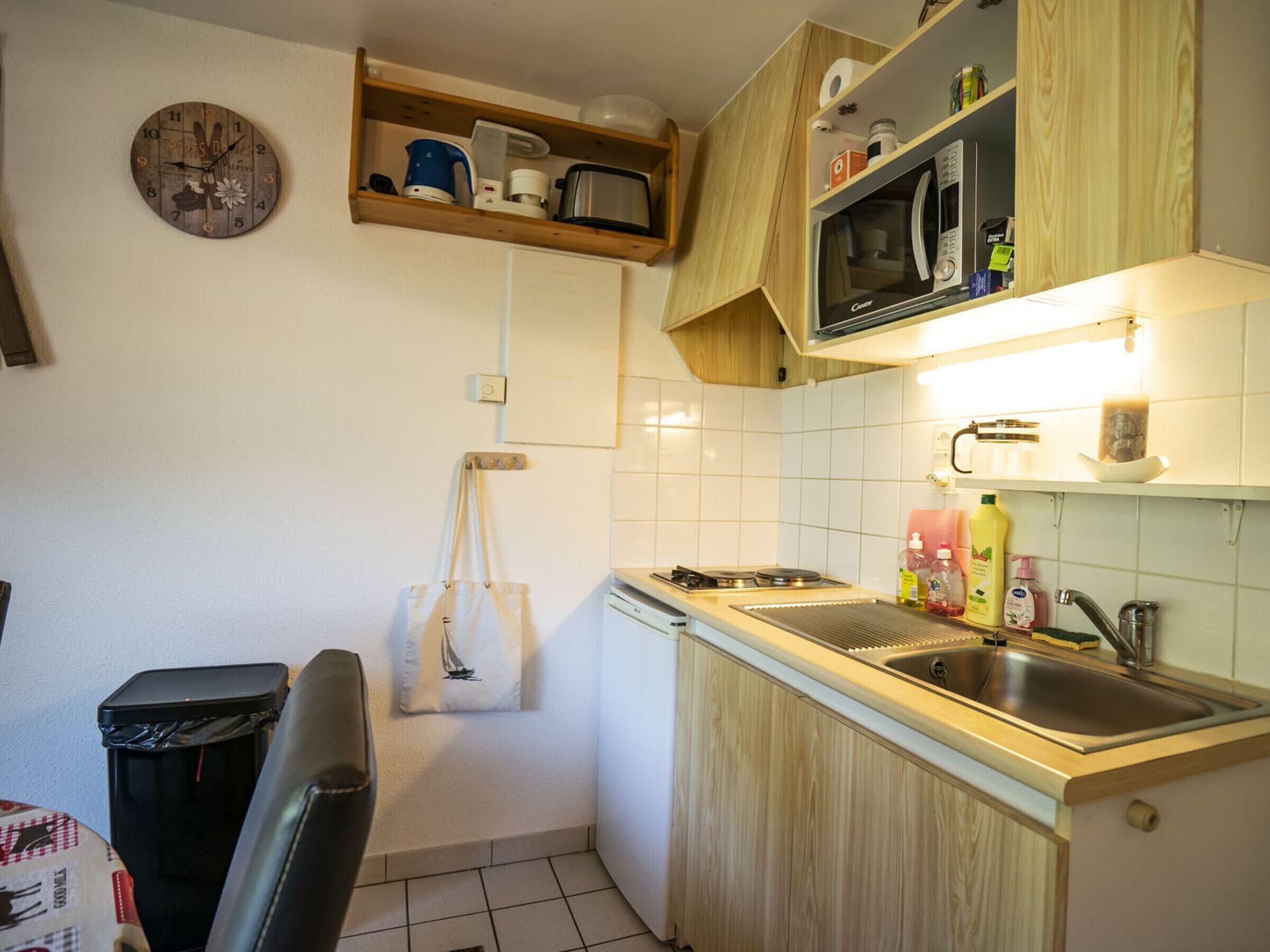 A compact kitchen with wooden cabinets, a microwave, a sink, and a clock on the wall.