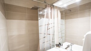 Un baño moderno con baldosas color beige, cabina de ducha de cristal, lavabo blanco con artículos de aseo y un espejo.