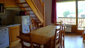 A kitchen with wooden cabinets, a white refrigerator, a stove, and a dining table with chairs. There is a staircase leading to a balcony with a view of the outside. - Calm Duplex T4 with parking, pets allowed in Samoëns (Samoëns)