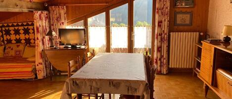 A cozy cabin-style room with a wooden ceiling, a dining table with chairs, a television on a wooden cabinet, and large windows with curtains.
