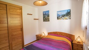 Une chambre avec une armoire en bois, un lit recouvert d’une couverture à rayures colorées, deux tableaux accrochés au mur et une table de chevet avec une lampe.