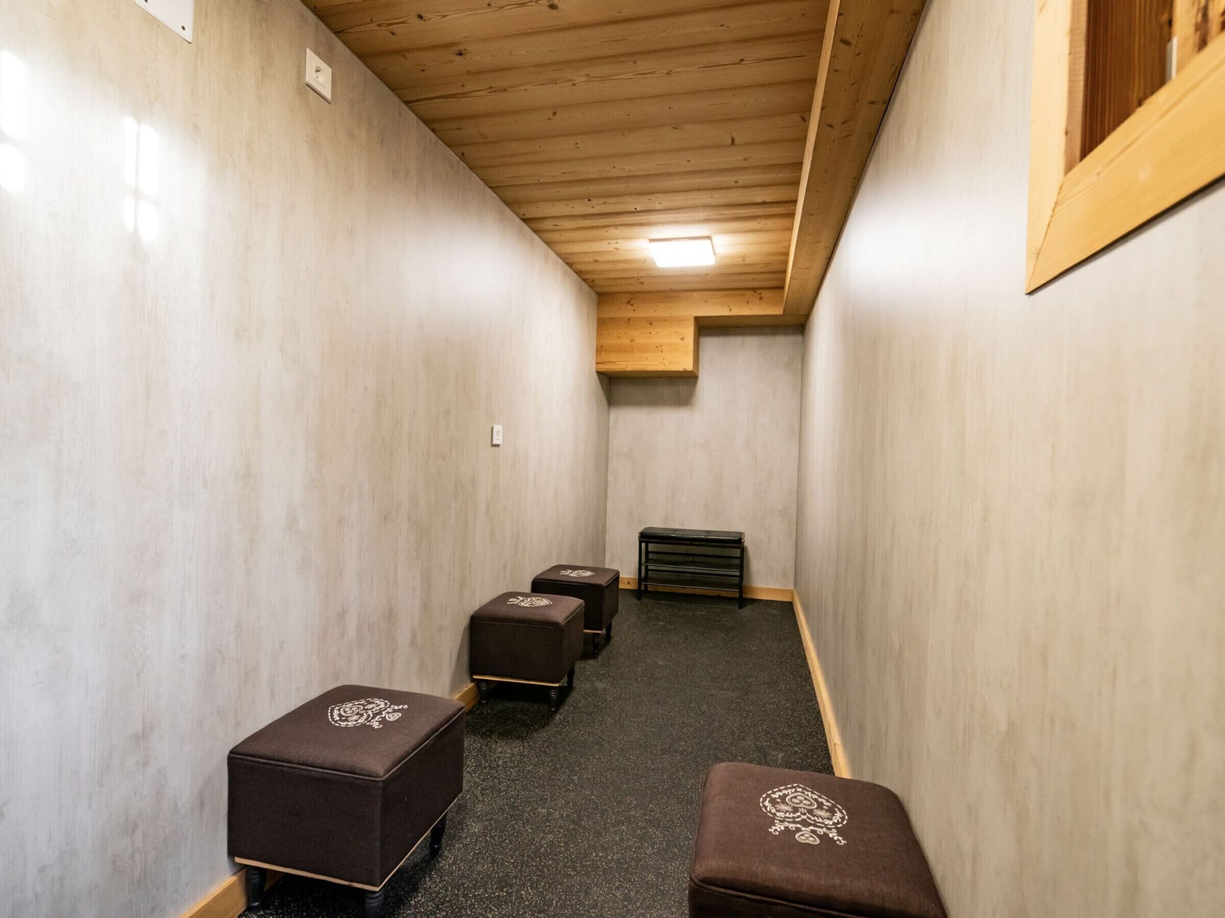 Een smalle gang met vier bruine poefs langs de muur, een houten plafond en een klein raam aan het einde.