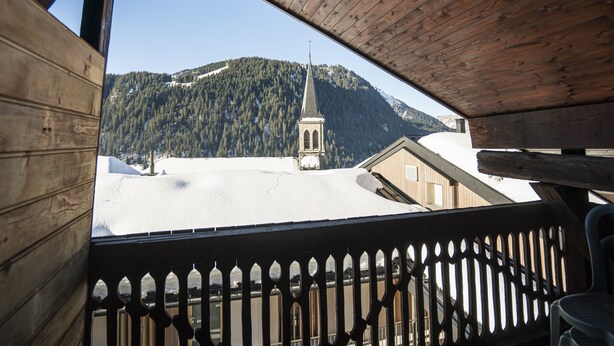 Un balcon en bois offrant une vue sur un paysage enneigé et sur une église dotée d’un clocher imposant.