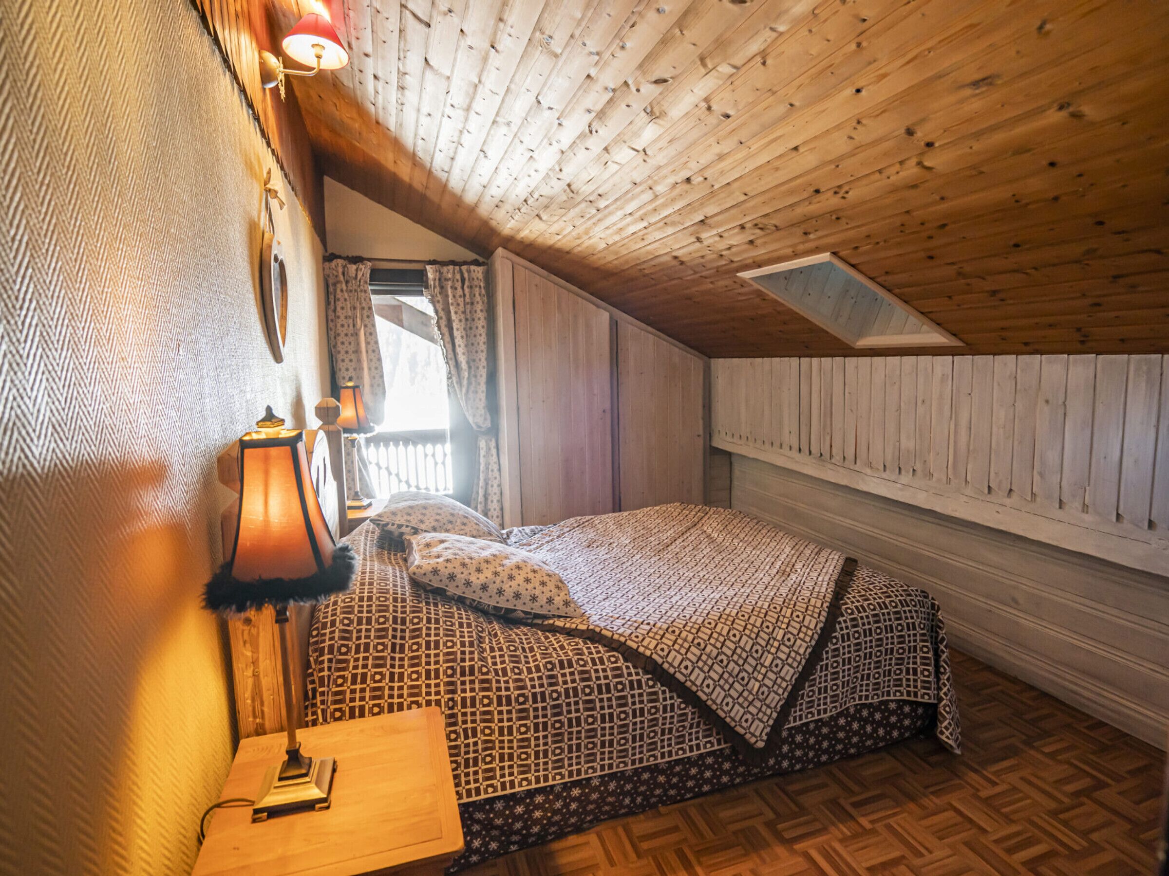 A cozy attic bedroom with a wooden ceiling, a bed with patterned bedding, a nightstand, and a lamp.