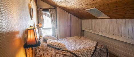 A cozy attic bedroom with a wooden ceiling, a bed with patterned bedding, a nightstand, and a lamp.